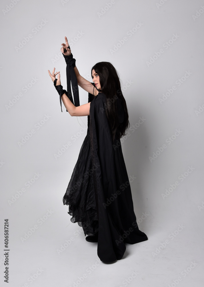 Full length portrait of pretty black haired woman wearing long dark gown and a cloak. Standing pose facing away from the camera, against a studio background. Full length portrait of pretty black haired woman wearing long dark gown and a cloak. Standing pose facing away from the camera, against a studio background.