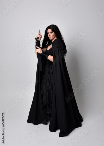 Full length portrait of pretty black haired woman wearing long dark gown and a cloak.  Standing pose facing away from the camera, against a  studio background.