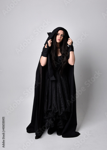 Full length portrait of pretty black haired woman wearing long dark gown and a cloak. Standing pose facing away from the camera, against a studio background.