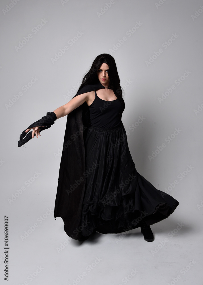 Naklejka premium Full length portrait of pretty black haired woman wearing long dark gown and a cloak. Standing pose facing away from the camera, against a studio background.