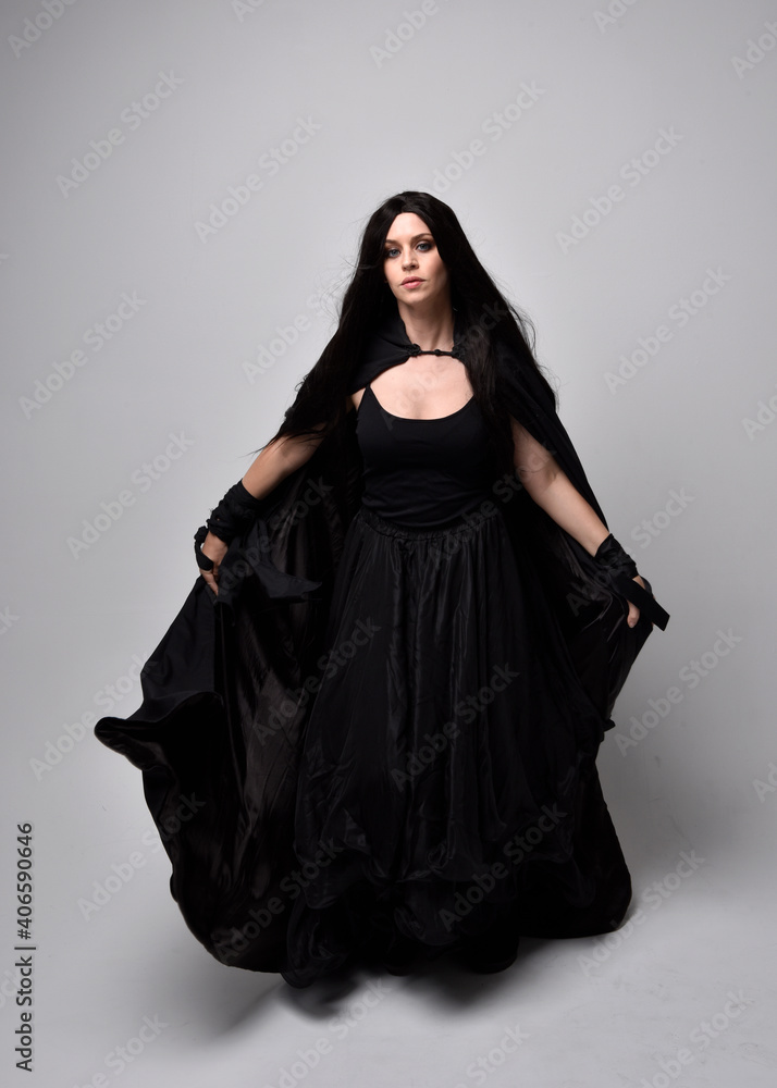 Full length portrait of pretty black haired woman wearing long dark gown and a cloak. Standing pose facing away from the camera, against a studio background. Full length portrait of pretty black haired woman wearing long dark gown and a cloak. Standing pose facing away from the camera, against a studio background.