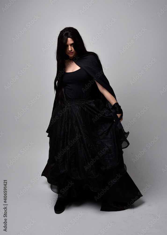 Full length portrait of pretty black haired woman wearing long dark gown and a cloak.  Standing pose facing away from the camera, against a  studio background.