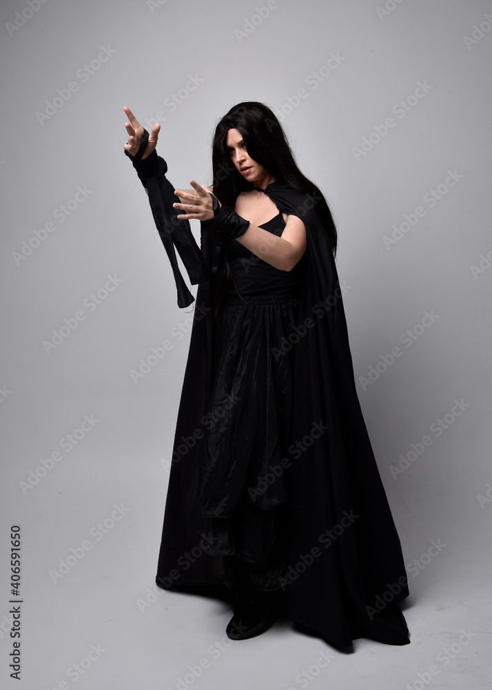 Full length portrait of pretty black haired woman wearing long dark gown and a cloak. Standing pose facing away from the camera, against a studio background. Full length portrait of pretty black haired woman wearing long dark gown and a cloak. Standing pose facing away from the camera, against a studio background.