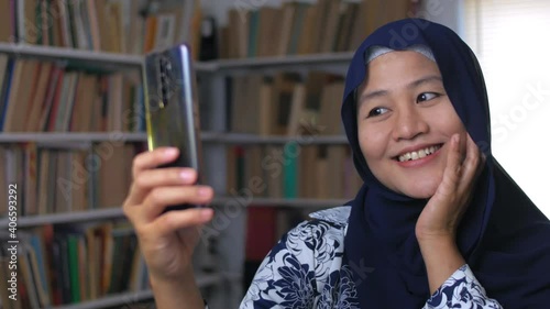 Happy beautiful Asian muslim woman taking selfie portrait with her smert phone in library photo