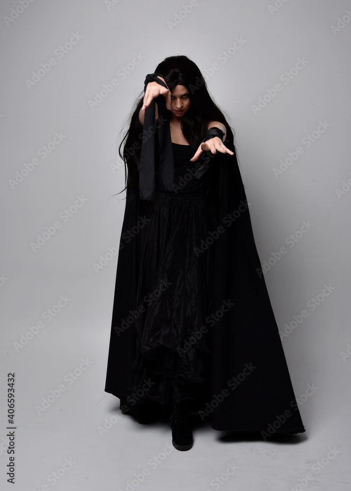 Full length portrait of pretty black haired woman wearing long dark gown and a cloak.  Standing pose facing away from the camera, against a  studio background.