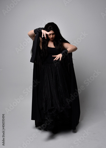 Full length portrait of pretty black haired woman wearing long dark gown and a cloak. Standing pose facing away from the camera, against a studio background.