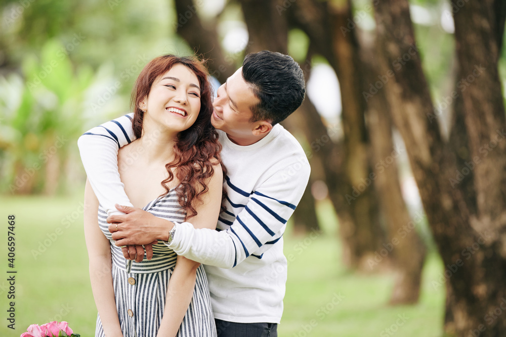 Happy Vietnamese young man hugging his girlfriend from behind and kissing her on cheek Happy Vietnamese young man hugging his girlfriend from behind and kissing her on cheek