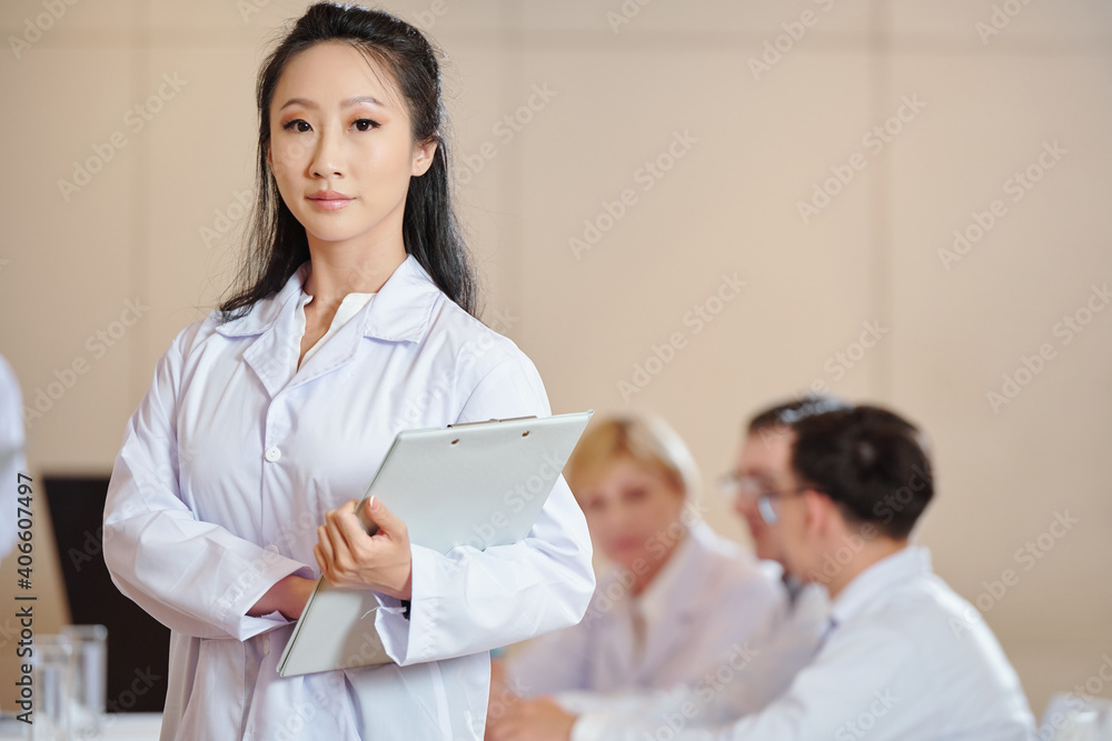 Serious young female Chinese scientist with clipboard standing in conference room and looking at camera Serious young female Chinese scientist with clipboard standing in conference room and looking at camera