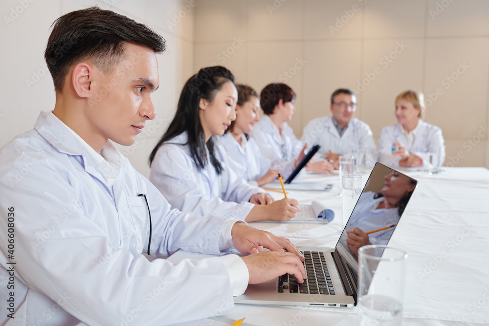 Serious young researcher working on laptop when attending meeting with scientists devoted to new coronavirus vaccine testing Serious young researcher working on laptop when attending meeting with scientists devoted to new coronavirus vaccine testing