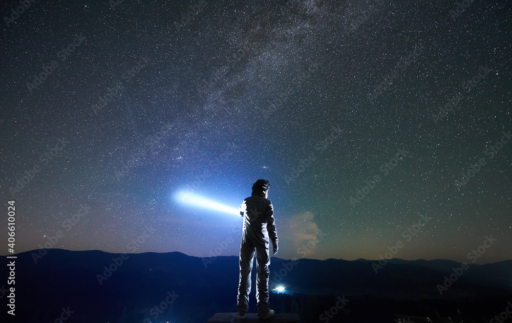 Cosmonaut wearing white space suit and helmet directs a blue ray of light into starry sky above horizon in the night, standing with his back to the camera in the mountains, space travel concept