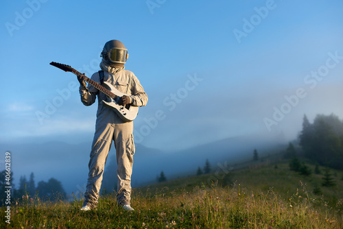 Astronaut wearing white space suit and helmet playing white guitar, standing on sunny green mountain glade in morning. Foggy hills, blue sky on background. Concept of astronautics, music and nature.