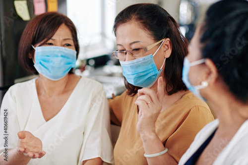 Group of senior Asian women in medical masks meeting at home due to coronavirus pandemic and discussing news and gossips