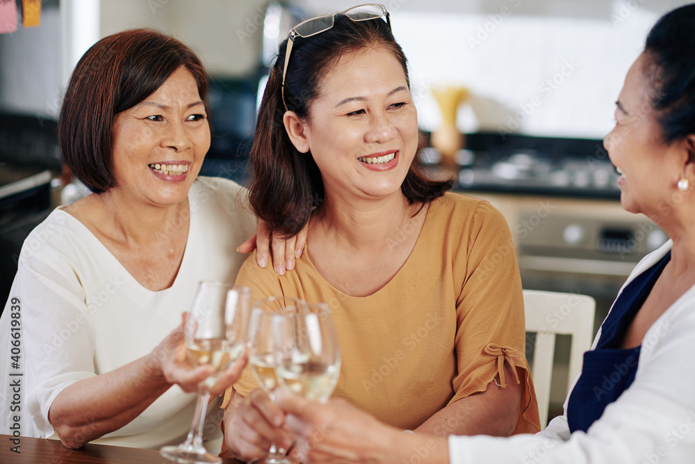 Group of cheerful Asian senior women drinking white wine at kitchen table and talking about health and family issues Group of cheerful Asian senior women drinking white wine at kitchen table and talking about health and family issues