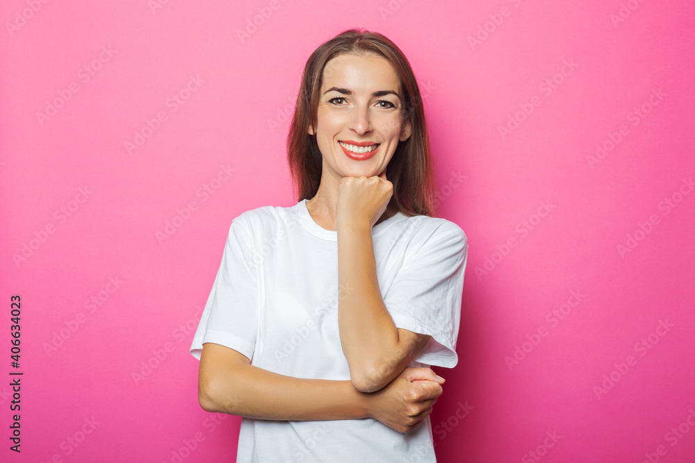 Fototapeta premium Young woman in white t-shirt holds hand under the chin, pink isolated background
