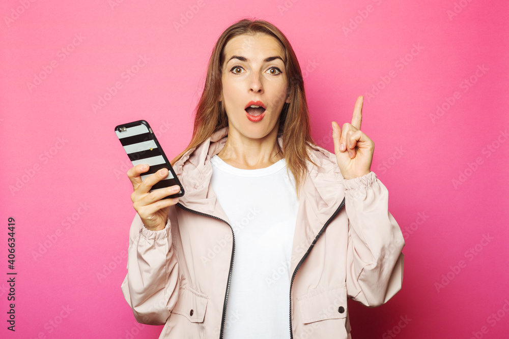 Young surprised woman holding a phone, pink isolated background