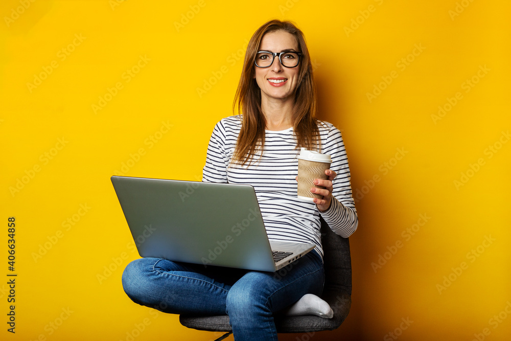 Young woman holding a cup of coffee, sitting on a chair, working on a laptop on an isolated yellow background Young woman holding a cup of coffee, sitting on a chair, working on a laptop on an isolated yellow background