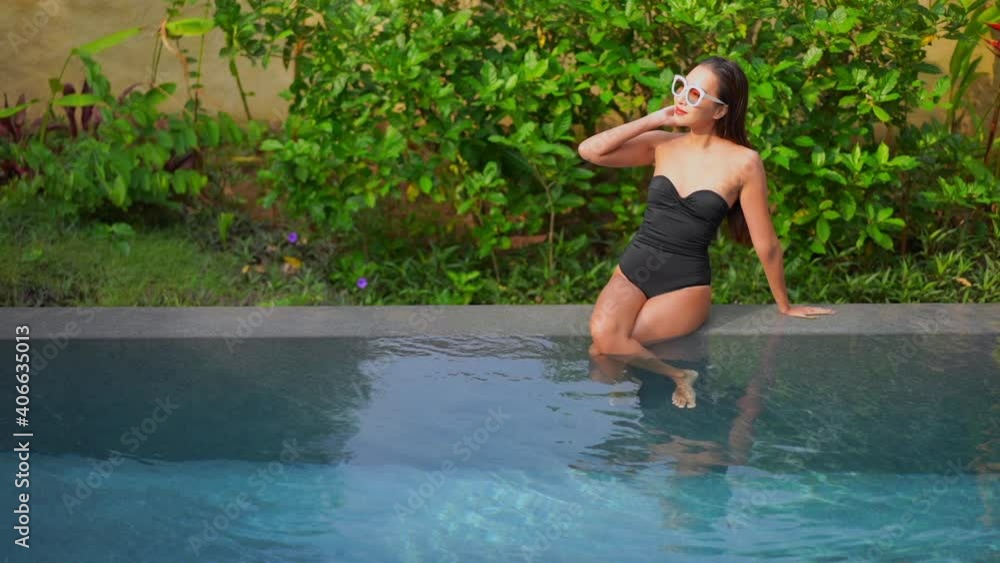Attractive Asian Woman in Swimsuit Sitting on Pool Edge on Tropical Destination. Slow Motion Attractive Asian Woman in Swimsuit Sitting on Pool Edge on Tropical Destination. Slow Motion
