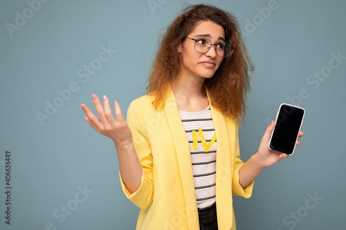 attractive asking young woman with curly dark blond hair wearing yellow jacket and optical glasses isolated on background holding and showing mobile phone with empty space for cutout looking to the