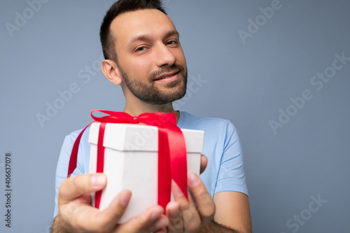 Portrait shot of handsome happy brunette unshaven young male person isolated over blue background wall wearing blue t-shirt holding white gift box with red ribbon and looking at camera