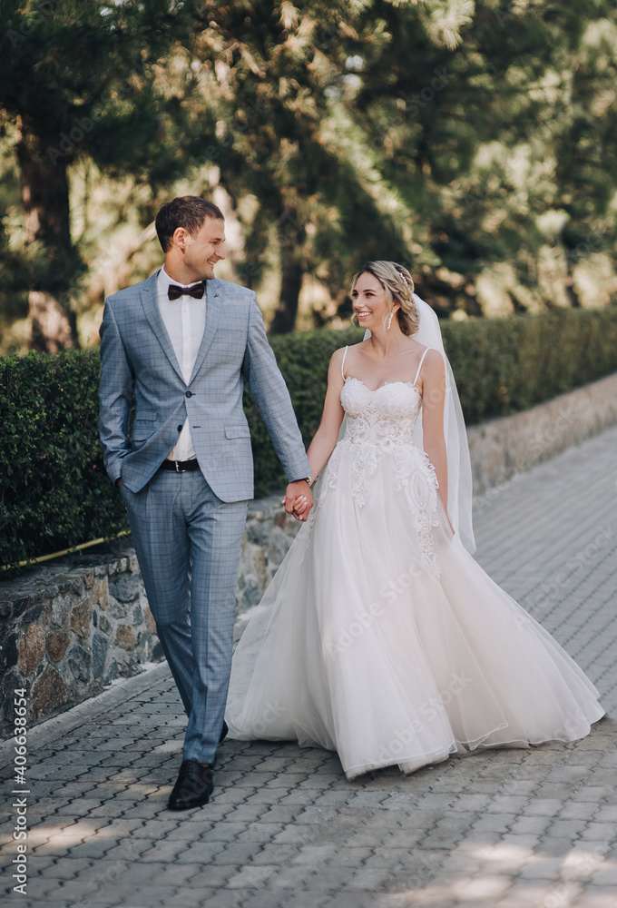 The groom in a gray checkered suit and the blonde bride in a white lace dress walk holding hands in the park on the boulevard. Wedding portrait of newlyweds in love