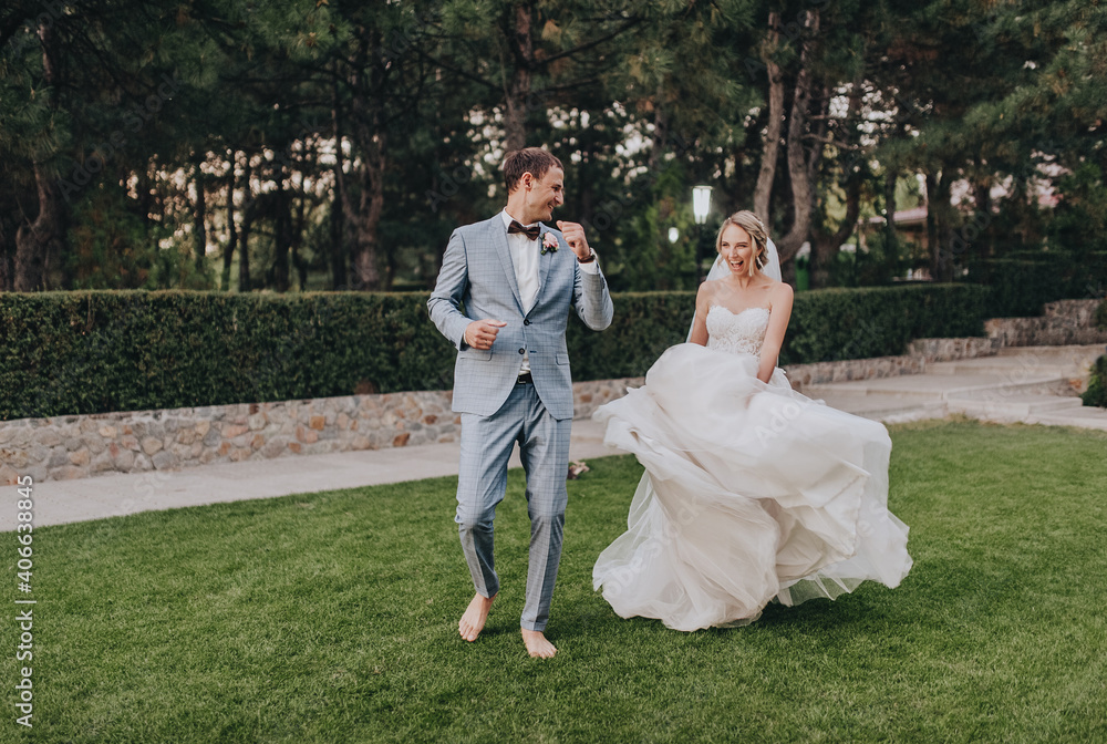Stylish groom in a gray checkered suit and a blonde bride in a long white dress cheerfully and funny dance in a clearing with green grass, against the background of a forest barefoot. Stylish groom in a gray checkered suit and a blonde bride in a long white dress cheerfully and funny dance in a clearing with green grass, against the background of a forest barefoot.