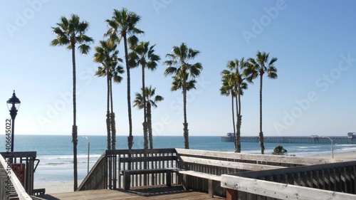 Fototapeta Naklejka Na Ścianę i Meble -  Wooden stairs, beach access in Oceanside, California USA. Coastal stairway, pacific ocean waves and palm trees. Vacations by sea in United States. Sunny tropical day, summertime aesthetic. Staircase.