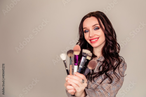 Young beautiful woman holding makeup brushes in her hands and posing on grey background at studio.