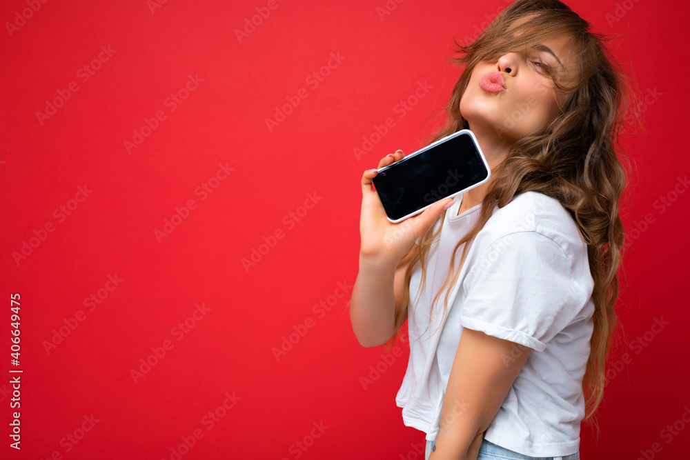 Photo of beautiful young blonde woman good looking wearing white t-shirt standing isolated on red background with copy space holding phone showing smartphone in hand with empty screen display for Photo of beautiful young blonde woman good looking wearing white t-shirt standing isolated on red background with copy space holding phone showing smartphone in hand with empty screen display for