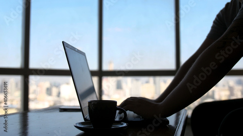 A concentrated young businessman is working on a laptop computer using his notebook sitting by the window with urban view and drinks coffee