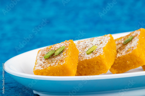 Traditional Indian Mithai Moong Dal Burfi Or Meetha Mung Daal Barfi Barfee Cooked In Desi Ghee Arranged In White Tray Viewed Low Angle From The Side On Bright Turquoise Blue Background