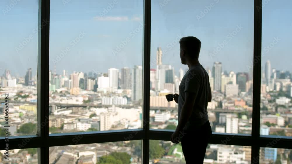 Dreaming european man stands by the window with great views and drinks hot coffee or tea, enjoying beautiful morning feeling happy Dreaming european man stands by the window with great views and drinks hot coffee or tea, enjoying beautiful morning feeling happy