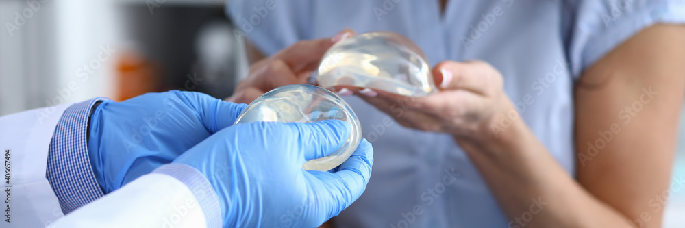 Fototapeta premium Patient of clinic hold breast implant in his hand and touch it with her finger. Doctor hold silicone material in his hand and show woman.