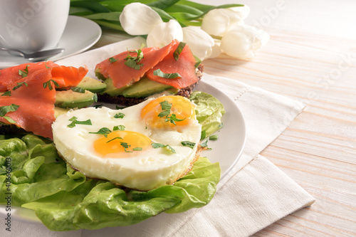 Holiday breakfast with love, fried eggs in heart shape, sandwiches with avocado and smoked salmon on lettuce, white tulips on a light painted wooden table, copy space
