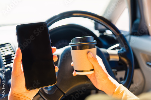 Asian woman drinking hot coffee takeaway cup inside a car and using smartphone blank screen while driving the car in the morning during going to work on highway, Transportation and vehicle concept © sorapop