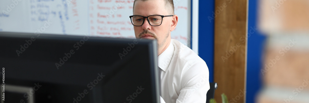 Serious man in glasses and shirt sit and look at computer monitor. In background is white board with text.