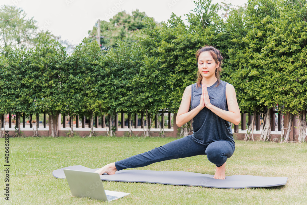 Fototapeta premium Asian young woman using laptop during online video training practicing yoga outdoors in meditate pose sitting on green grass in nature a field garden park, Stretching health care concept