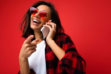 Closeup photo of pretty happy young brunet woman wearing stylish red shirt white t-shirt and red sunglasses isolated over red background communicating on mobile phone looking at camera