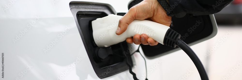 Male hand insert electric white gun into car. White electric car be recharged at charging station. Male hand insert electric white gun into car. White electric car be recharged at charging station.