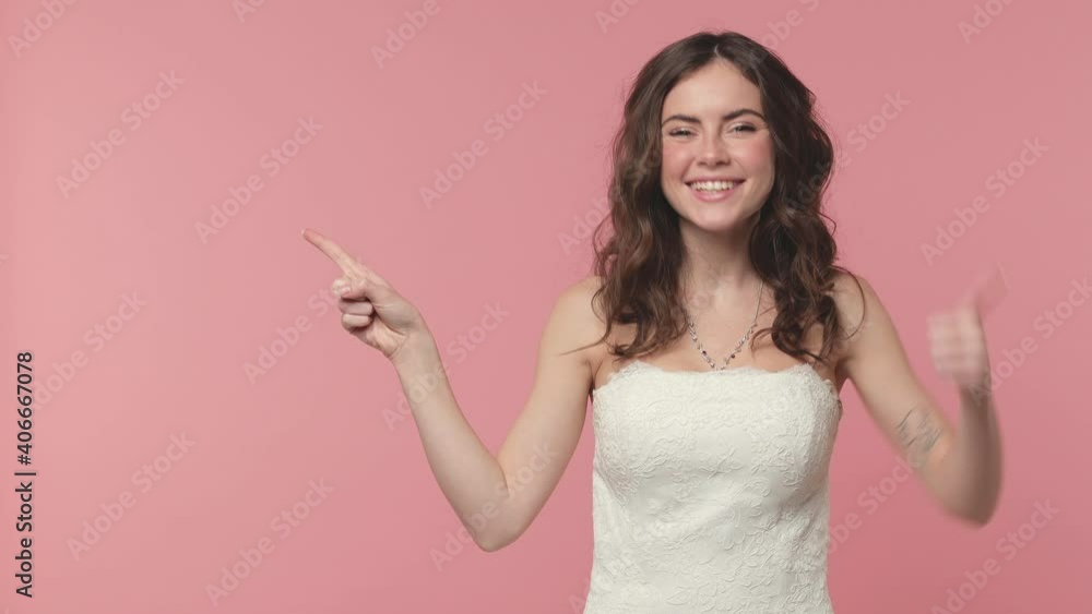 Shocked excited bride young brunette woman 20s in beautiful white wedding dress posing isolated on pink background studio. Wedding concept. Pointing finger aside say wow showing thumb up like gesture Shocked excited bride young brunette woman 20s in beautiful white wedding dress posing isolated on pink background studio. Wedding concept. Pointing finger aside say wow showing thumb up like gesture