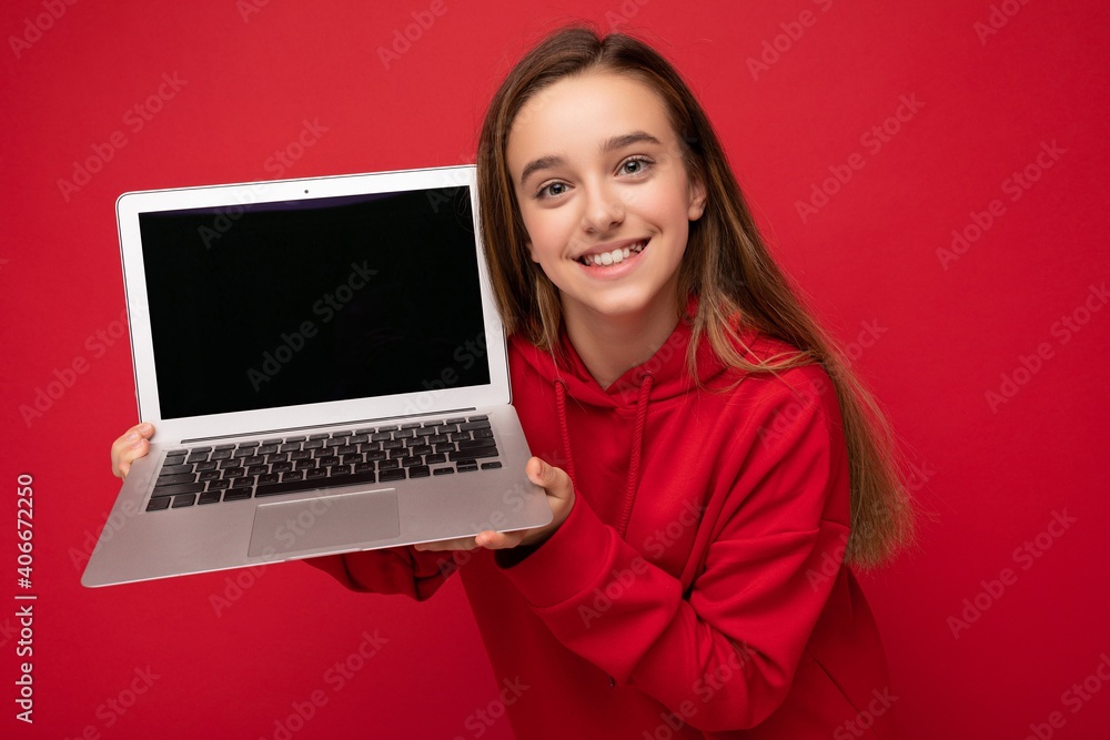 Close-up portrait photo of beautiful happy smiling girl with long hair wearing red hoodie holding computer laptop looking at camera isolated over red wall background Close-up portrait photo of beautiful happy smiling girl with long hair wearing red hoodie holding computer laptop looking at camera isolated over red wall background