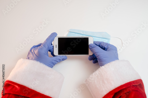 Santa Claus hands holding smart phone and a medical mask over white table background.
