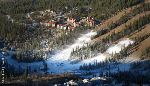 Beschneiung (technische Schneeerzeugung) in einem Skigebiet mit Skiliften und Skipisten. Im Hintergrund eine Hotelanlage. CO2 Fußabdruck im Kontext von Klimawandel und globaler Erderwärmung.
