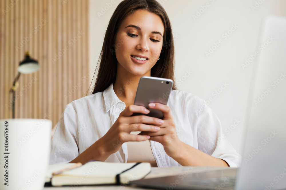 Attractive business woman sits at table in front of laptop and talks on mobile phone, negotiates on the phone. Beautiful brunette girl uses gadgets for work. Online training, remote work from home Attractive business woman sits at table in front of laptop and talks on mobile phone, negotiates on the phone. Beautiful brunette girl uses gadgets for work. Online training, remote work from home