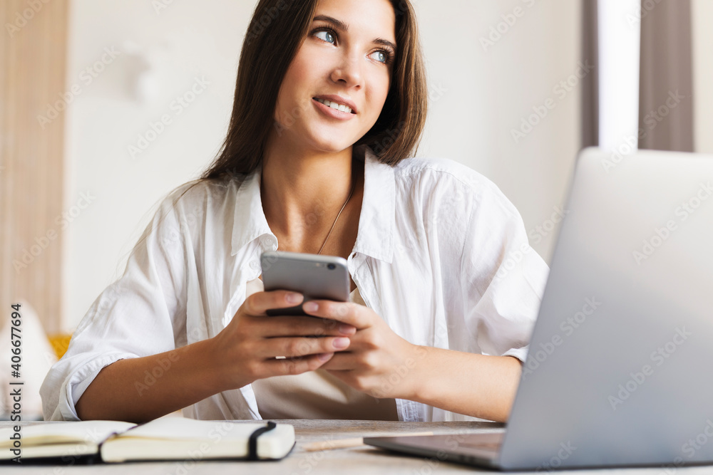 Attractive business woman sits at table in front of laptop and talks on mobile phone, negotiates on the phone. Beautiful brunette girl uses gadgets for work. Online training, remote work from home Attractive business woman sits at table in front of laptop and talks on mobile phone, negotiates on the phone. Beautiful brunette girl uses gadgets for work. Online training, remote work from home