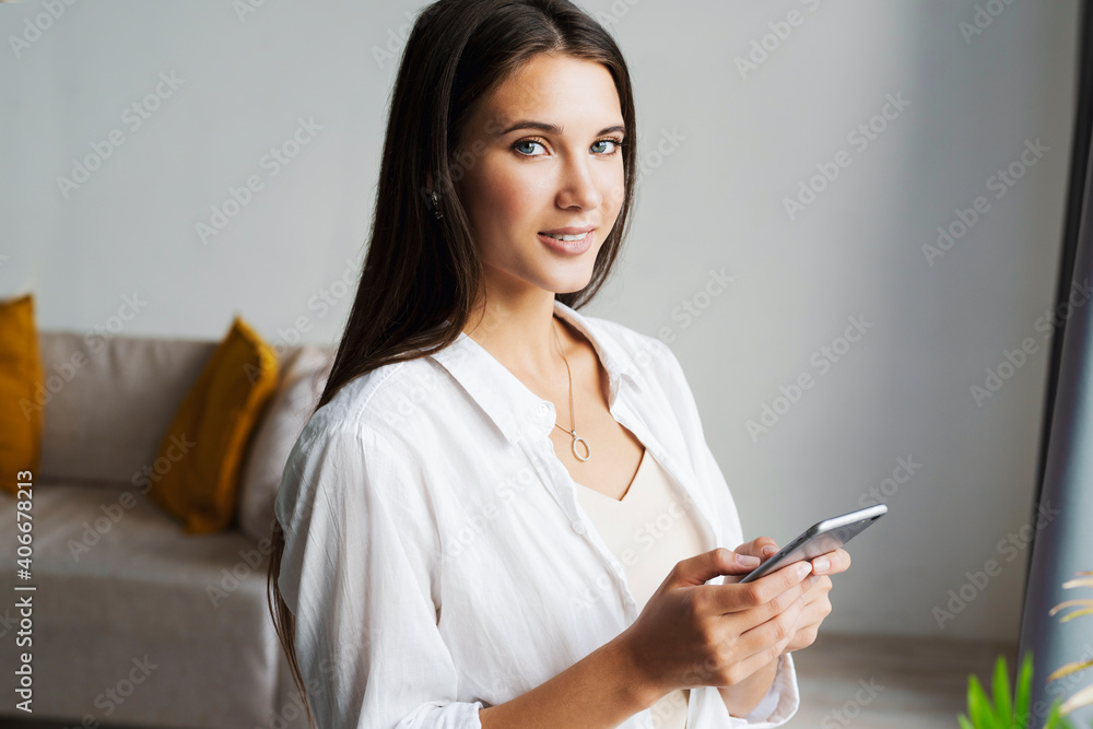 Portrait of business woman, holds smartphone in her hands. Beautiful girl uses mobile phone for work, calls clients, sends text messages. Millennial girl in white shirt stands against white wall Portrait of business woman, holds smartphone in her hands. Beautiful girl uses mobile phone for work, calls clients, sends text messages. Millennial girl in white shirt stands against white wall