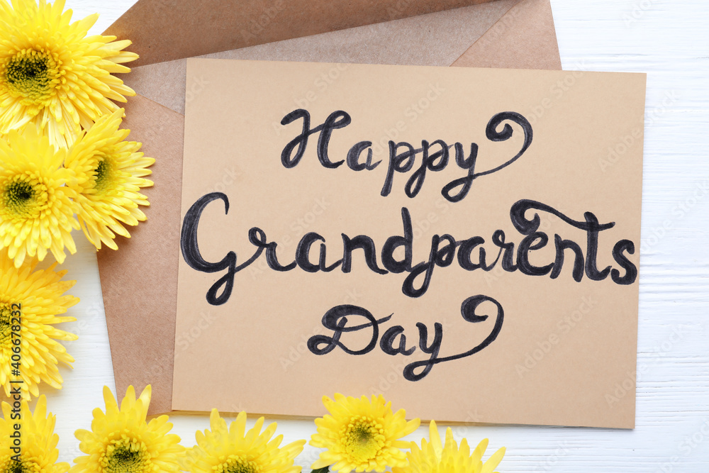 Naklejka premium Beautiful yellow flowers, envelope and card with phrase Happy Grandparents Day on white wooden table, flat lay