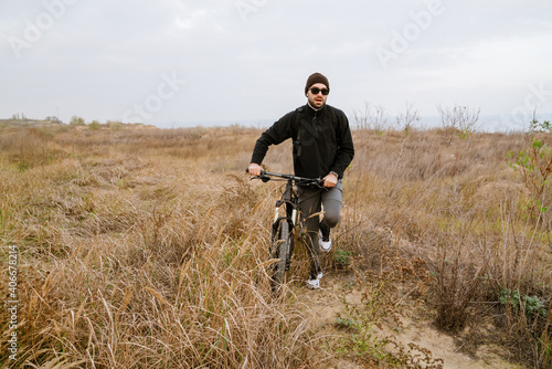 Athletic unshaven guy in sunglasses walking with his bicycle on field