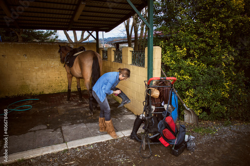 Woman cleaning her riding boots with her brown horse in the background