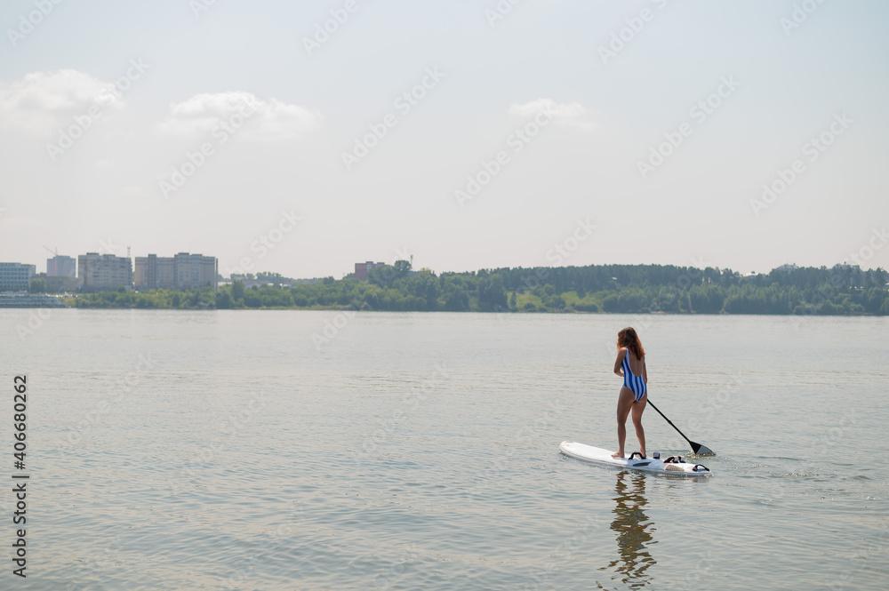 Naklejka premium Caucasian woman in a striped swimsuit rides on a SUP board. The girl prefers active rest.