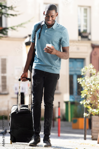 Full body young black man walking with luggage and mobile phone on city street